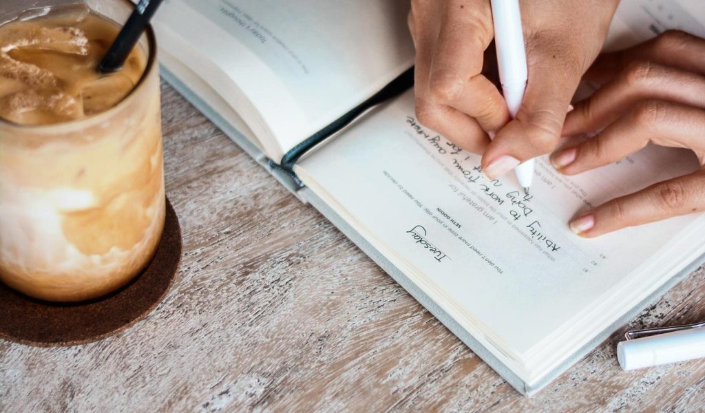 Person writing in a gratitude notebook with an iced coffee